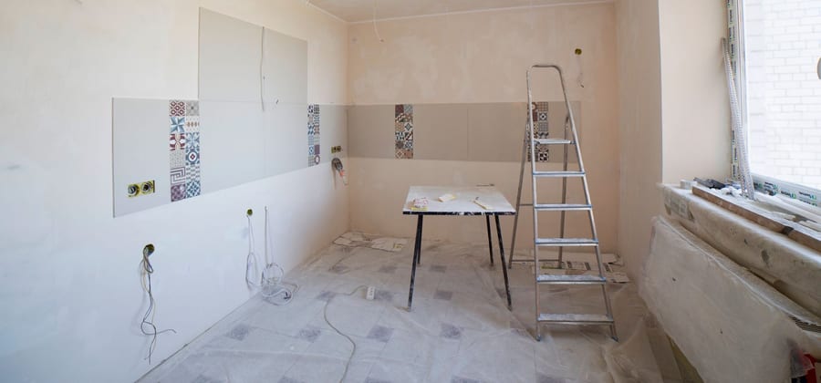 Ett kök mitt i renoveringen: nyspacklade väggar, kakel på väg upp och en stege mitt i rummet.
